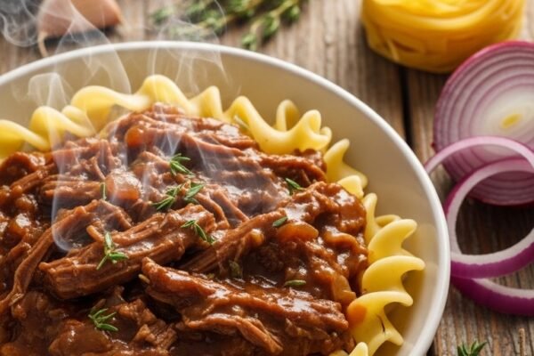 A steaming bowl of homemade beef and noodles recipe with rich brown gravy, tender shredded beef, and egg noodles served on a rustic wooden table with warm golden lighting.