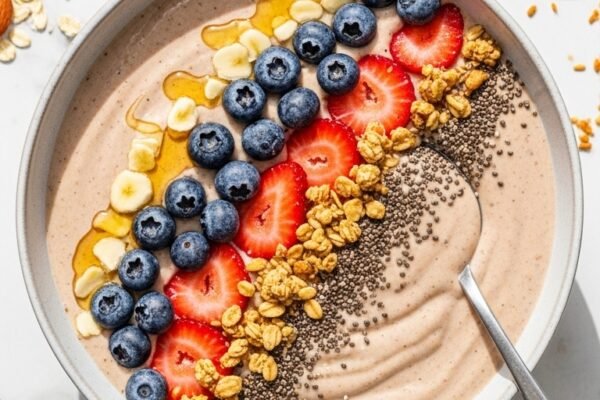 A perfect thick smoothie recipe in a wide ceramic bowl with creamy spoonable texture topped with fresh blueberries sliced strawberries granola honey drizzle and chia seeds on a white marble surface surrounded by frozen fruits in bright natural light.