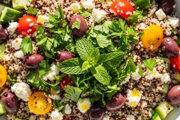 Vibrant quinoa salad in a large white ceramic bowl with fluffy quinoa grains, diced cucumber, cherry tomatoes, kalamata olives, crumbled feta and fresh herbs with golden olive oil drizzle on white marble in bright natural light