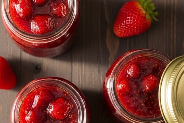 Three glass mason jars of vibrant homemade strawberry jam with golden lids surrounded by fresh strawberries and a rustic wooden spoon with jam dripping on a dark wooden table in warm natural light