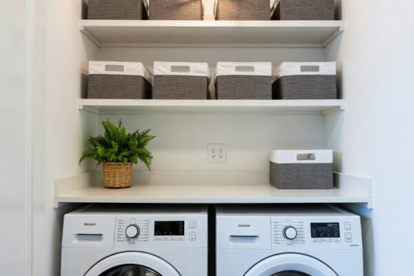 A beautifully transformed modern laundry room featuring white floor-to-ceiling cabinets, a deep utility sink, folding station countertop above front-load washer and dryer, open shelving with organized baskets, and bright layered lighting — showcasing the most practical and stylish laundry room ideas for a complete chore space transformation.