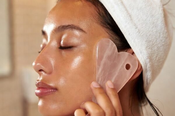 A beautifully serene beauty lifestyle photograph of a woman with naturally radiant and sculpted glowing skin gently using a smooth rose quartz gua sha tool along her cheekbone and jawline demonstrating an effective and calming gua sha facial routine for natural lymphatic drainage skin sculpting and effortless everyday radiance without harsh tools.