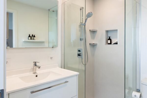 A beautifully transformed small full bathroom featuring light neutral walls a large wall mirror wall mounted vanity corner shower with clear glass door floating shelves and smart space saving fixtures creating a bright open and stylish tiny bathroom in bright natural light.