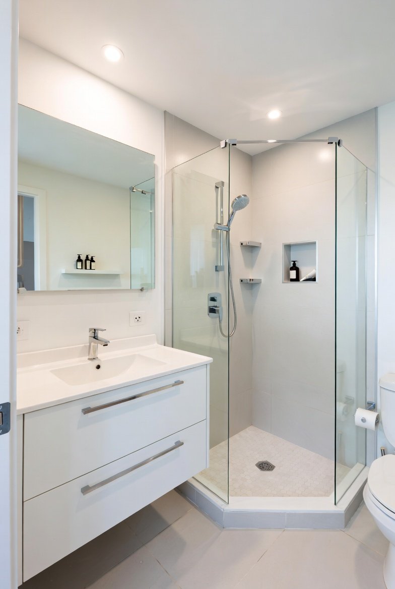 A beautifully transformed small full bathroom featuring light neutral walls a large wall mirror wall mounted vanity corner shower with clear glass door floating shelves and smart space saving fixtures creating a bright open and stylish tiny bathroom in bright natural light.